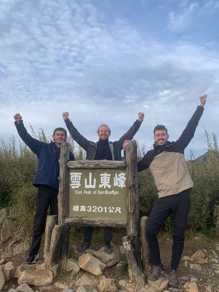 Trois randonneurs bras levés devant le panneau du East Peak of Xue Mountain à 3201 mètres d'altitude